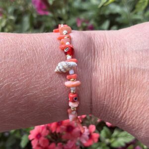 Close-up van een pols met de "Tara" armband uit de zomercollectie van Little Caribbean. De armband heeft kleurrijke koraalrode en oranje kralen, gecombineerd met kleine gouden accenten en een schelpbedeltje, perfect voor een zomerse look. Op de achtergrond zijn kleurrijke bloemen zichtbaar.