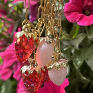 Close-up van de "Pink Strawberry's" ketting uit de zomercollectie van Little Caribbean, met charmante aardbeienbedeltjes in roze en rood, aan een gouden ketting. Op de achtergrond zijn levendige roze bloemen zichtbaar.