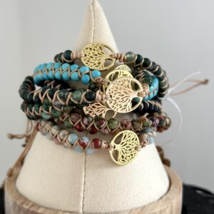 Stapel van "Beach Serenity's" armbanden van Little Caribbean, met kleurrijke turquoise, groene en aardse kralen en gouden levensboom bedels, op een decoratieve standaard.