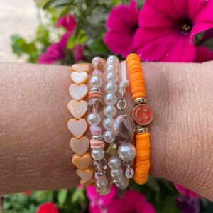 Set van vijf "Beach Vibes" armbanden van Little Caribbean met oranje, perzikkleurige, parelwitte en roze kralen, versierd met hartjes en een smiley bedel, gedragen om de pols met kleurrijke bloemen op de achtergrond.