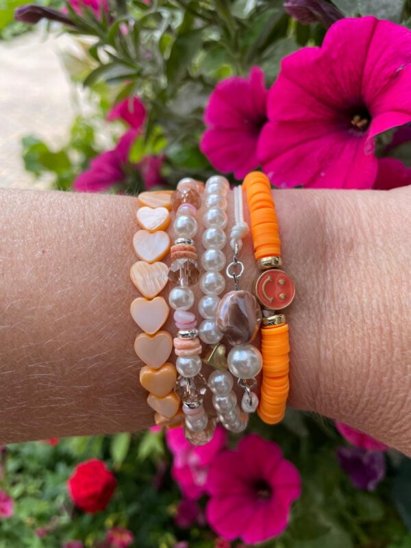 Set van vijf "Beach Vibes" armbanden van Little Caribbean met oranje, perzikkleurige, parelwitte en roze kralen, versierd met hartjes en een smiley bedel, gedragen om de pols met kleurrijke bloemen op de achtergrond.