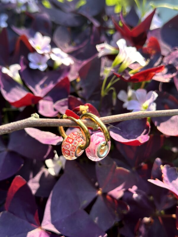 Paar "Bling Beaches Pink" oorbellen van Little Caribbean, met roze en rode kralen en goudkleurige accenten, hangend aan een tak met paarse bladeren en witte bloemen op de achtergrond.