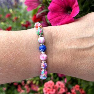 "Bloom Color" armband van Little Caribbean, met levendige roze, blauwe, groene, rode en witte kralen met bloemenpatronen en zilverkleurige accenten, gedragen om de pols met kleurrijke bloemen op de achtergrond.