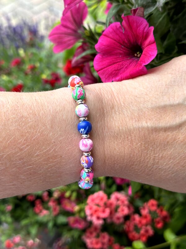 "Bloom Color" armband van Little Caribbean, met levendige roze, blauwe, groene, rode en witte kralen met bloemenpatronen en zilverkleurige accenten, gedragen om de pols met kleurrijke bloemen op de achtergrond.