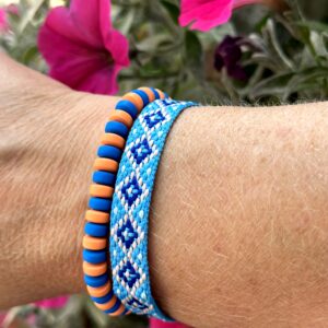 Set van twee armbanden genaamd "Blue Orange" van Little Caribbean, met een handgeweven armband in blauw, wit en roze, en een kralenarmband in oranje en blauw, gedragen om de pols met kleurrijke bloemen op de achtergrond.