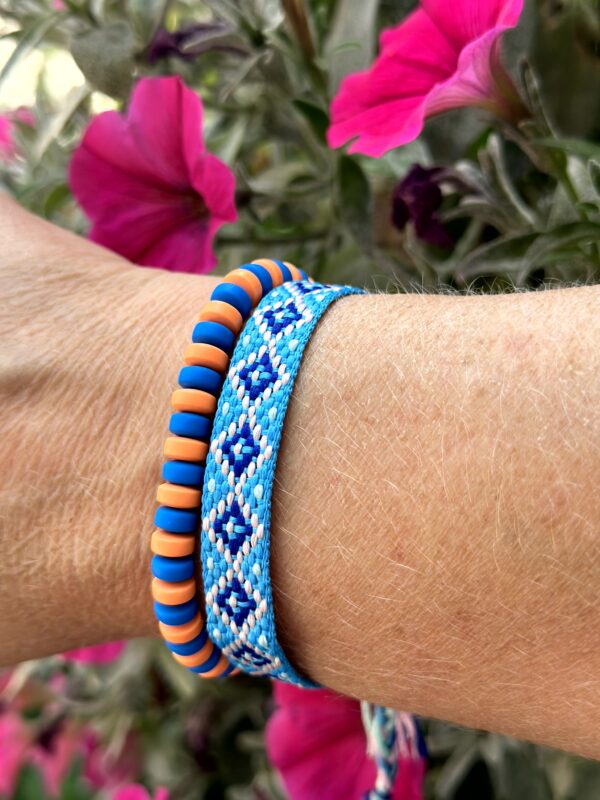 Set van twee armbanden genaamd "Blue Orange" van Little Caribbean, met een handgeweven armband in blauw, wit en roze, en een kralenarmband in oranje en blauw, gedragen om de pols met kleurrijke bloemen op de achtergrond.