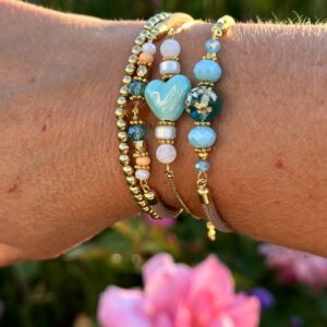 Set van drie "Blue Paradise" armbanden van Little Caribbean, met blauwe, turquoise, witte en gouden kralen, gedragen om de pols met een kleurrijke bloem op de achtergrond.