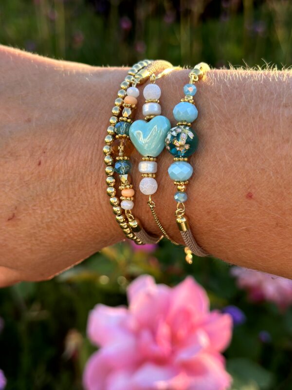 Set van drie "Blue Paradise" armbanden van Little Caribbean, met blauwe, turquoise, witte en gouden kralen, gedragen om de pols met een kleurrijke bloem op de achtergrond.