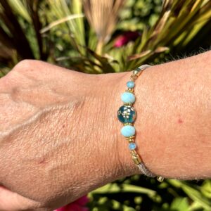 "Blue Paradise" armband van Little Caribbean, met blauwe en turquoise kralen en goudkleurige accenten, gedragen om de pols met een groene en kleurrijke tuin op de achtergrond.