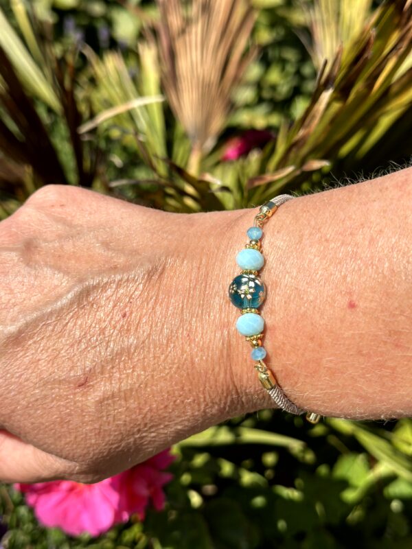 "Blue Paradise" armband van Little Caribbean, met blauwe en turquoise kralen en goudkleurige accenten, gedragen om de pols met een groene en kleurrijke tuin op de achtergrond.