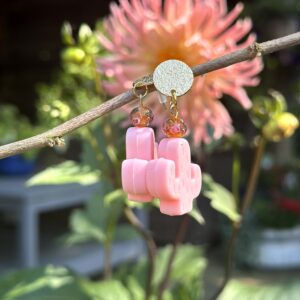 Paar "Caribbean Pink Cactus" oorbellen van Little Caribbean, met roze cactusvormen, glinsterende oranje kralen en goudkleurige accenten, hangend aan een tak met een kleurrijke bloem op de achtergrond.