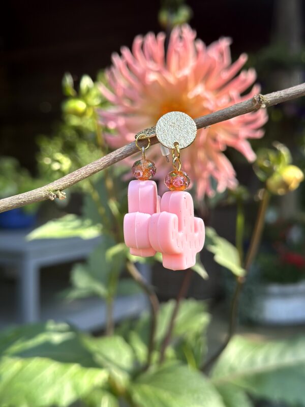 Paar "Caribbean Pink Cactus" oorbellen van Little Caribbean, met roze cactusvormen, glinsterende oranje kralen en goudkleurige accenten, hangend aan een tak met een kleurrijke bloem op de achtergrond.