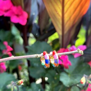 Paar "Happy Arubas" oorbellen van Little Caribbean, met kleurrijke rood, blauw, groen en oranje vlakken en goudkleurige accenten, hangend aan een tak met kleurrijke bloemen op de achtergrond.