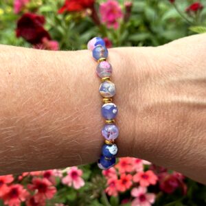 "Island Bliss" armband van Little Caribbean, met blauwe, roze en paarse kralen en goudkleurige accenten, gedragen om de pols met kleurrijke bloemen op de achtergrond.