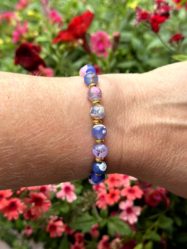 "Island Bliss" armband van Little Caribbean, met blauwe, roze en paarse kralen en goudkleurige accenten, gedragen om de pols met kleurrijke bloemen op de achtergrond.