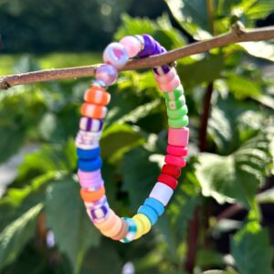 "Beach Colors" kinderarmbandje van Little Caribbean, met een regenboog van kleurrijke kralen en speelse patronen, hangend aan een tak met groene bladeren op de achtergrond.