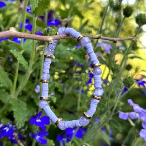 "Purple Ibiza" kinderarmbandje van Little Caribbean, met paarse kraaltjes en goudkleurige accenten, hangend aan een tak met paarse bloemen op de achtergrond.