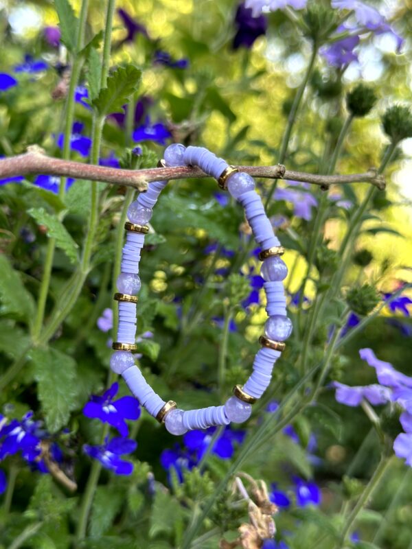 "Purple Ibiza" kinderarmbandje van Little Caribbean, met paarse kraaltjes en goudkleurige accenten, hangend aan een tak met paarse bloemen op de achtergrond.