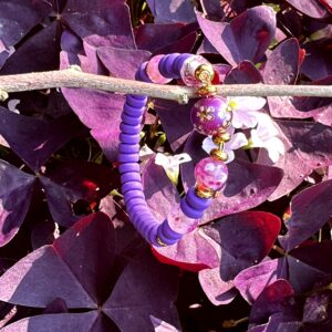 "Purple Sweet" kinderarmbandje van Little Caribbean, met paarse en roze kralen en goudkleurige accenten, hangend aan een tak met paarse bladeren op de achtergrond.