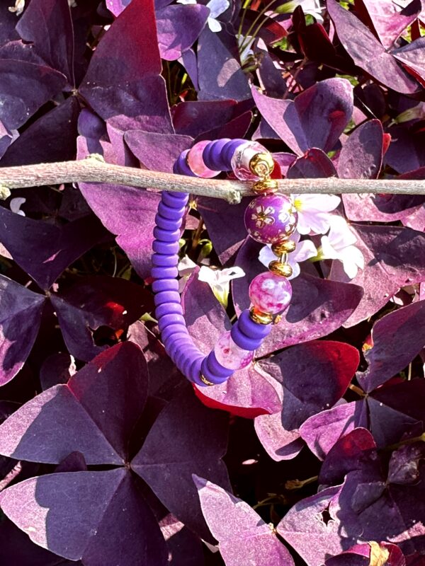 "Purple Sweet" kinderarmbandje van Little Caribbean, met paarse en roze kralen en goudkleurige accenten, hangend aan een tak met paarse bladeren op de achtergrond.