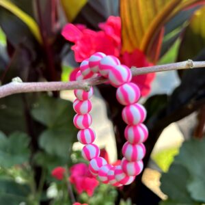 "Sweet Candy Pink" kinderarmbandje van Little Caribbean, met roze en witte kralen die eruitzien als snoepjes, hangend aan een tak met kleurrijke bloemen op de achtergrond.