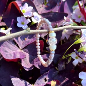 "Zeemeermin" kinderarmbandje van Little Caribbean, met zachte paarse kralen, goudkleurige accenten en een glinsterende kraal, hangend aan een tak met paarse bladeren en bloemen op de achtergrond.