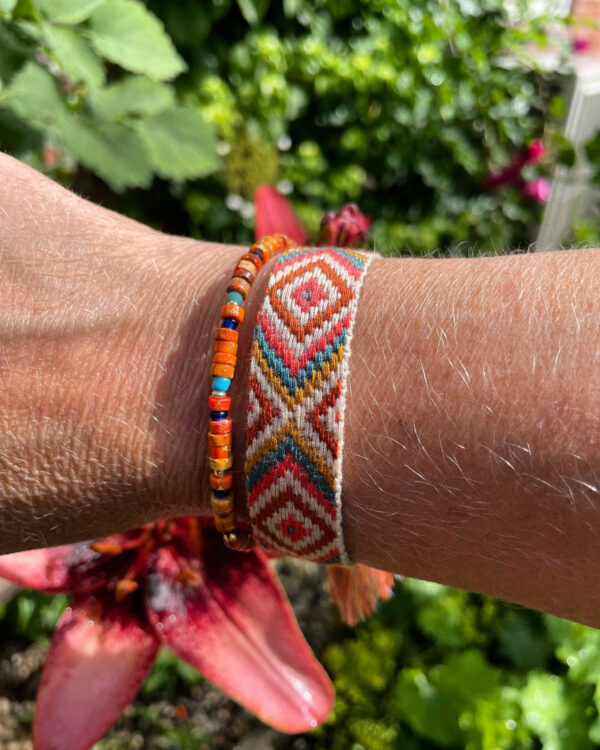 "Ocean Harmony" armbandenset van Little Caribbean, met een geweven armband met traditionele patronen en een kralenarmband in warme tinten van oranje, rood en turquoise, gedragen aan een pols met groene bladeren op de achtergrond.