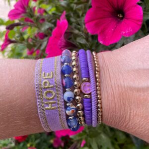 "Ocean Jewel Purple" armbandenset van Little Caribbean, met paarse, gouden en blauwe kralen, een "HOPE" armband, gedragen aan een pols met kleurrijke bloemen op de achtergrond.