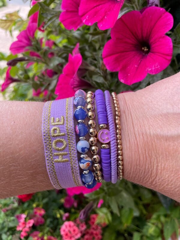 "Ocean Jewel Purple" armbandenset van Little Caribbean, met paarse, gouden en blauwe kralen, een "HOPE" armband, gedragen aan een pols met kleurrijke bloemen op de achtergrond.