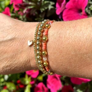 "Ocean Orange Treasure" armbandenset van Little Caribbean, met oranje en gouden kralen, een hartjesbedel, gedragen aan een pols met kleurrijke bloemen op de achtergrond.