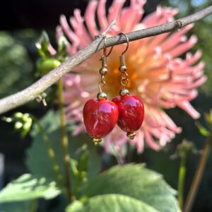 "Red Valentines" oorbellen van Little Caribbean, met felrode hartvormige bedels en gouden accenten, hangend aan een tak met een bloem op de achtergrond.