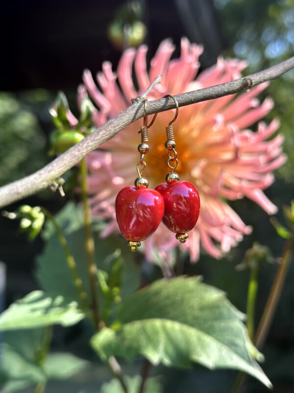 "Red Valentines" oorbellen van Little Caribbean, met felrode hartvormige bedels en gouden accenten, hangend aan een tak met een bloem op de achtergrond.