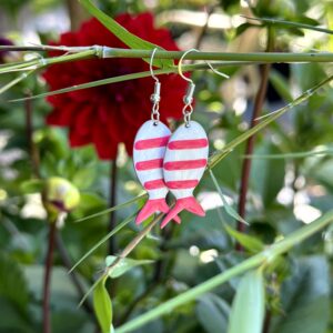 "Pink Vis" oorbellen van Little Caribbean, met roze en witte strepen, gefotografeerd tussen groene bladeren en een bloem.