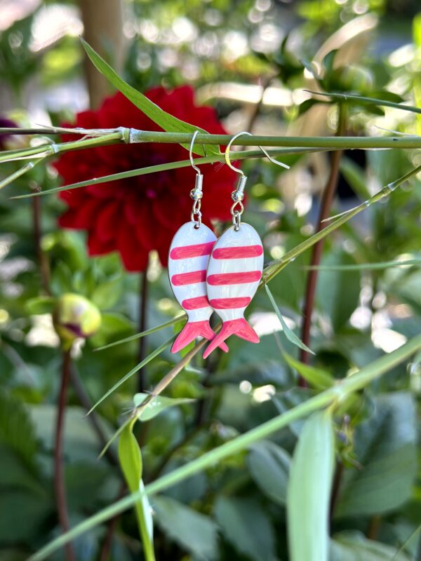 "Pink Vis" oorbellen van Little Caribbean, met roze en witte strepen, gefotografeerd tussen groene bladeren en een bloem.