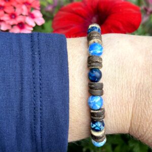 "Palm Blue" armband van Little Caribbean, met houten kralen en natuurstenen in blauwtinten, gedragen op een pols met kleurrijke bloemen op de achtergrond.