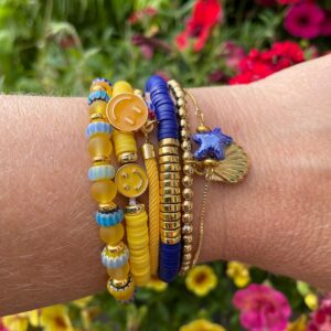 "Sunset Glow" armbanden set van Little Caribbean met vrolijke smiley-kralen, gouden accenten en charmante schelp en sterretje, gedragen op een pols.