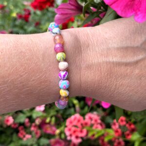 "Tropical Lagoon" armband van Little Caribbean met kralen in roze, blauw, geel en groen, gedragen door een arm met een kleurrijke bloemenachtergrond.