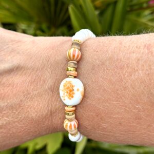 "Tropical Paradise" armband van Little Caribbean met witte, gouden en oranje kralen, gedragen door een arm met een groene achtergrond.